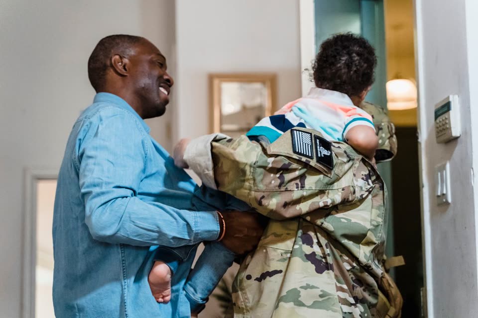 A service member in uniform carrying their child through the front door as their spouse smiles
